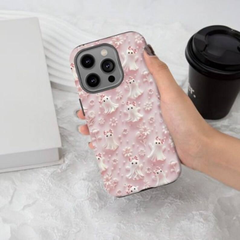 以下が含まれることがあります： ピンクの携帯電話ケースで、白い幽霊とピンクの花のパターンが描かれています。このケースはiPhone 14 Pro Max用に設計されています。