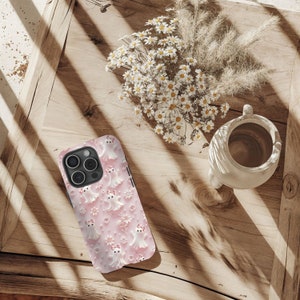 以下が含まれることがあります： 白い幽霊とピンクの花のパターンが施されたピンクのiPhoneケース。ケースはトリプルレンズカメラ付きのiPhone用に設計されています。
