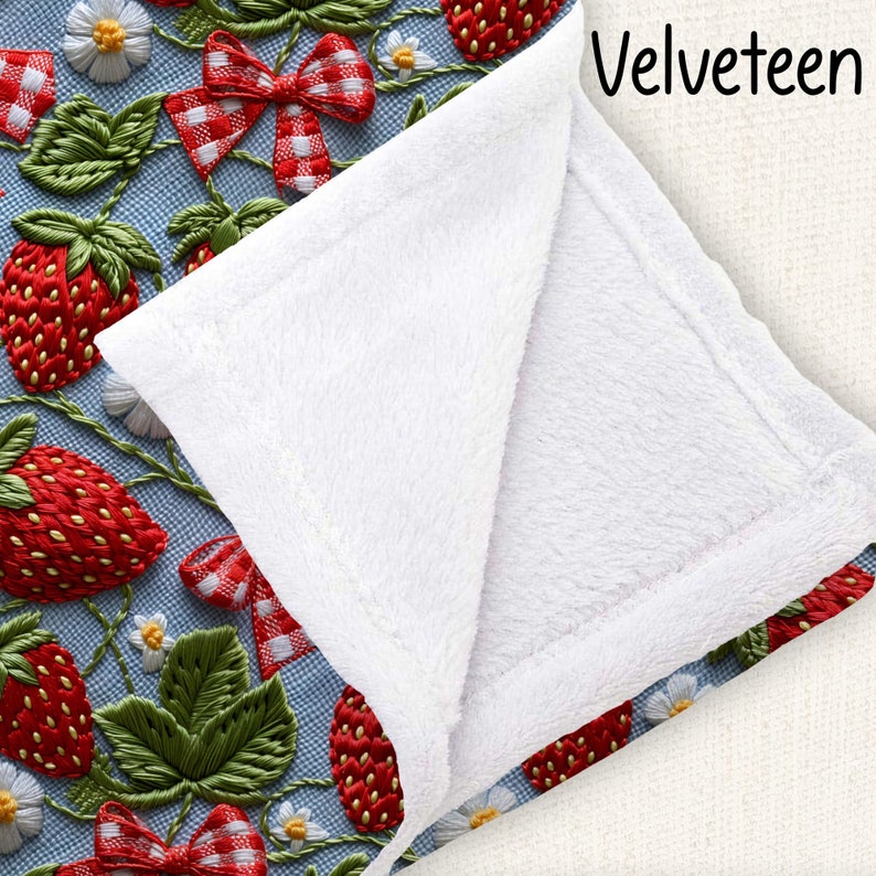 May include: A white throw blanket with a soft, plush texture. The blanket is folded in half and has a blue and white embroidered pattern of strawberries and bows on one side. The text "Velveteen" is printed on the other side of the blanket.