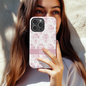 Op de afbeelding: Een roze en witte bloemen telefoonhoes met de gepersonaliseerde naam "Eli" gedrukt aan de onderkant. De hoes is ontworpen voor een iPhone met een camerabult.
