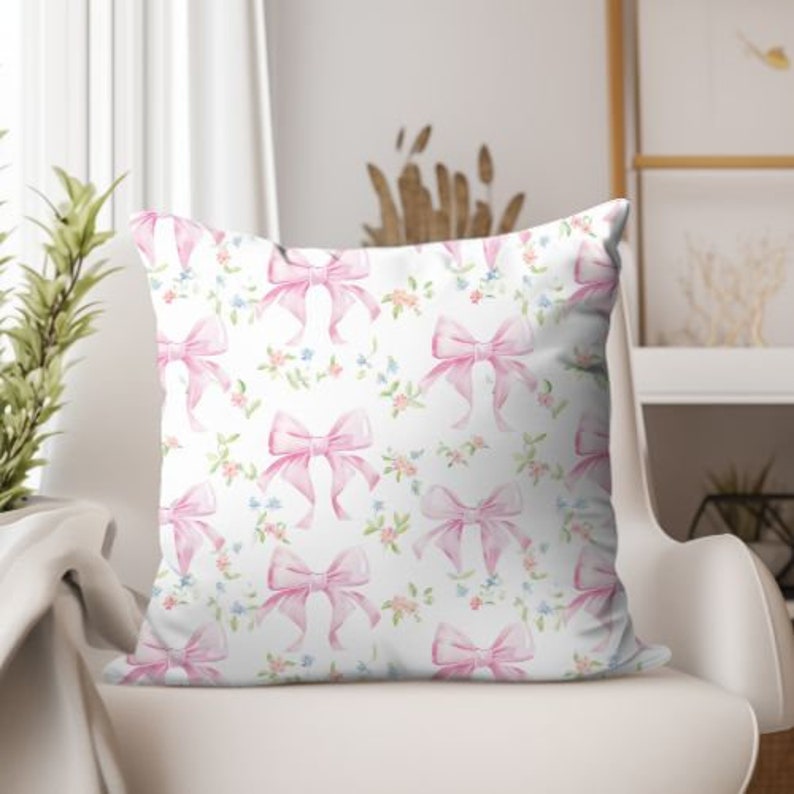 K&ouml;nnte beinhalten: Ein wei&szlig;es Kissen mit einem Muster aus rosa Schleifen. Die Schleifen sind im Aquarell-Stil gehalten und haben eine hellrosa Farbe. Der Hintergrund ist wei&szlig; mit kleinen rosa Bl&uuml;ten.