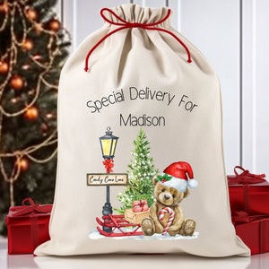 Può includere: Un sacco di Babbo Natale in tela bianca con laccetti rossi. Il sacco ha un messaggio personalizzato che dice "Consegna speciale per Madison". La parte anteriore del sacco presenta un'illustrazione ad acquerello di un orsacchiotto con un cappello di Babbo Natale, che tiene un bastoncino di zucchero e siede accanto a un lampione con un cartello che dice "Candy Cane Lane".
