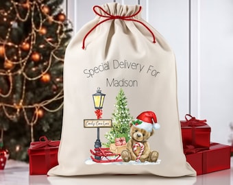 May include: A white canvas Santa sack with red drawstrings. The sack has a personalised message that says "Special Delivery For Madison". The front of the sack features a watercolour illustration of a teddy bear wearing a Santa hat, holding a candy cane, and sitting next to a street lamp with a sign that says "Candy Cane Lane".