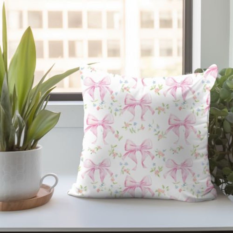 K&ouml;nnte beinhalten: Ein wei&szlig;es Kissen mit einem Muster aus rosa Schleifen. Die Schleifen sind mit Aquarellfarben bemalt und haben eine hellrosa und dunkelrosa Farbe. Der Hintergrund ist wei&szlig; mit kleinen blauen und rosa Blumen.