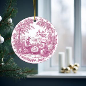 May include: A round Christmas ornament with a pink and white toile design. The illustration depicts a nativity scene with figures and an angel. The ornament is hanging from a gold string and is displayed on a Christmas tree.