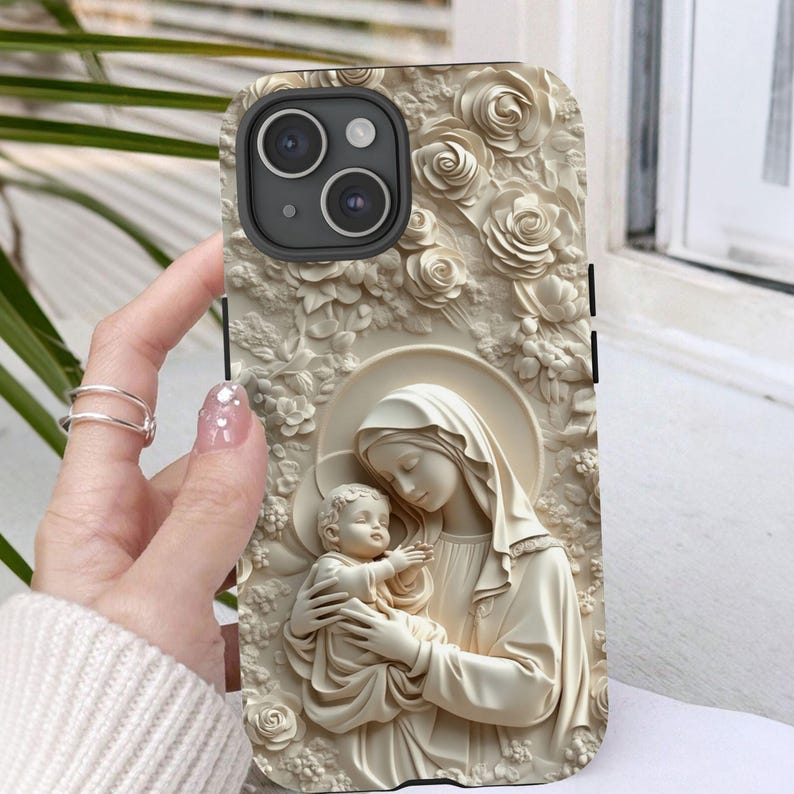 May include: A white phone case with a 3D design of the Virgin Mary holding a baby Jesus. The design is surrounded by white flowers.