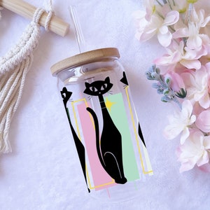 Puede incluir: Una botella de agua reutilizable de vidrio transparente con tapa de bambú y un gráfico en blanco y negro de gatos. El gráfico presenta tres gatos con un diseño retro y un fondo rosa, verde y amarillo.