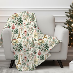 May include: A white throw blanket featuring a festive Parisian Christmas design. The pattern includes illustrations of the Eiffel Tower, houses, trees, and snowflakes.