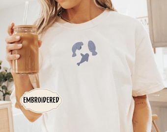 Peut inclure: T-shirt blanc avec un motif brodé de trois lamantins de couleur violet clair. Le texte "EMBROIDERED" est sur une étiquette blanche.