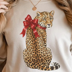 Puede incluir: Sudadera beige con una ilustración de leopardo con un lazo rojo. El leopardo tiene un pelaje manchado y está sentado erguido. El lazo es de color rojo vibrante y añade un toque festivo al diseño.
