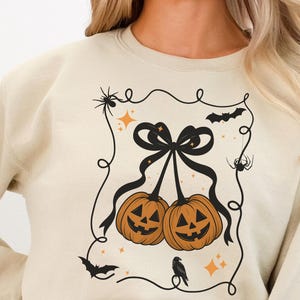 May include: A cream-coloured sweatshirt with a Halloween design. The graphic shows two orange pumpkins with black bows, surrounded by spiders, bats, and stars. A crow sits beneath the pumpkins, all within a decorative frame.