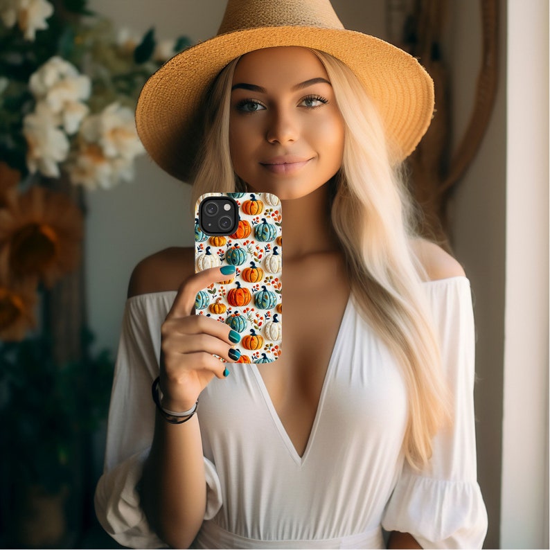 Peut inclure: Une coque de t&eacute;l&eacute;phone blanche avec un motif de citrouilles orange, blanches et bleues. La coque est tenue par une personne portant une chemise blanche et un chapeau de paille.
