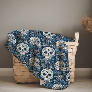 May include: A blue and white throw blanket with a sugar skull pattern in a woven wicker basket. The blanket features a repeating pattern of sugar skulls, flowers, and spiderwebs.