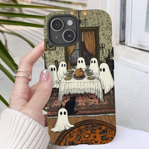 May include: A white iPhone case with a spooky Halloween design. The case features a group of ghosts gathered around a table with a turkey, surrounded by a black cat and a decorative rug.