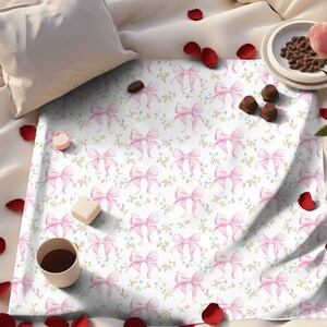 May include: A white fabric with a repeating pattern of pink bows and small flowers. The fabric is on a bed with rose petals scattered around it.