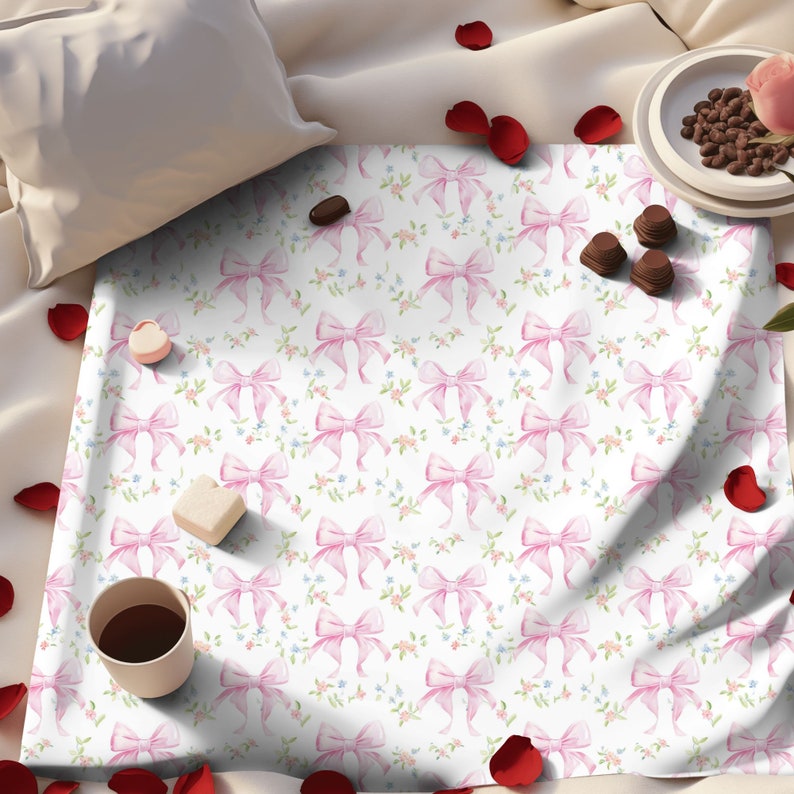 May include: A white fabric with a repeating pattern of pink bows and small flowers. The fabric is on a bed with rose petals scattered around it.