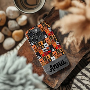 May include: A black phone case with a colorful patchwork design featuring Halloween icons like ghosts, bats, pumpkins, and cats. The case has a wood-look finish with the name "Anna" engraved on the bottom.