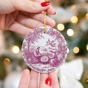 May include: A round Christmas ornament with a religious scene in a burgundy and white toile design. The ornament features a depiction of the Nativity with Mary, Joseph, and baby Jesus. A gold string is attached for hanging.