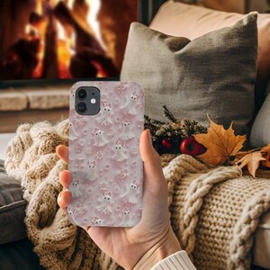 以下が含まれることがあります： 白い幽霊と花のパターンが施されたピンクの携帯電話ケース。ケースはiPhone用です。