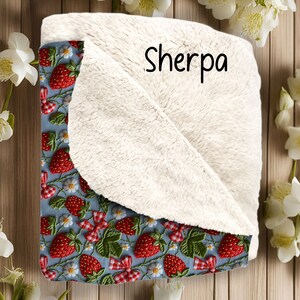 May include: A blue and white throw blanket with a sherpa lining. The blanket features a repeating pattern of embroidered strawberries and flowers. The text "Sherpa" is printed on the blanket.