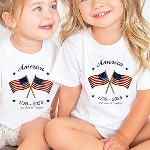 May include: Two children wearing white t-shirts with a patriotic design. The shirts feature the word "America" above crossed American flags and the text "1776-2026 250 Years of Freedom."