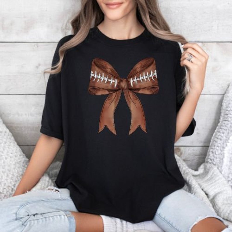 May include: A black t-shirt with a brown football-shaped bow on the front. The bow has white stitching details.