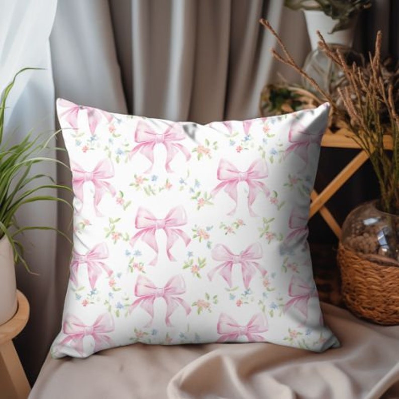 K&ouml;nnte beinhalten: Ein wei&szlig;es Kissen mit einem Muster aus rosa Schleifen. Die Schleifen sind im Aquarell-Stil gehalten und haben eine hellrosa Farbe. Der Hintergrund ist wei&szlig; mit kleinen floralen Details.