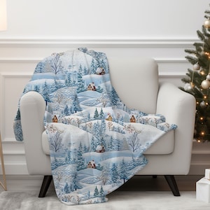 Può includere: Una morbida coperta blu e bianca con una scena invernale con case e alberi coperti di neve.