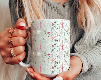 Può includere: Una tazza in ceramica bianca con un motivo floreale con fiori selvatici rosa, bianchi e verdi. La tazza è tenuta dalle mani di una persona.