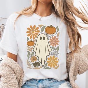 May include: White t-shirt featuring a retro-style Halloween design. The graphic includes a friendly ghost, pumpkins, flowers, spiderwebs, and a black cat in shades of orange, beige, and green. A fun, seasonal tee.