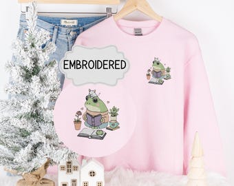 Può includere: Felpa rosa con un ricamo di una rana che indossa un cappello e legge un libro. Il testo "EMBROIDERED" è su un'etichetta bianca.