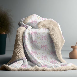 May include: A soft, plush throw blanket with a white background and a pink watercolor bow pattern. The blanket has a beige sherpa backing.