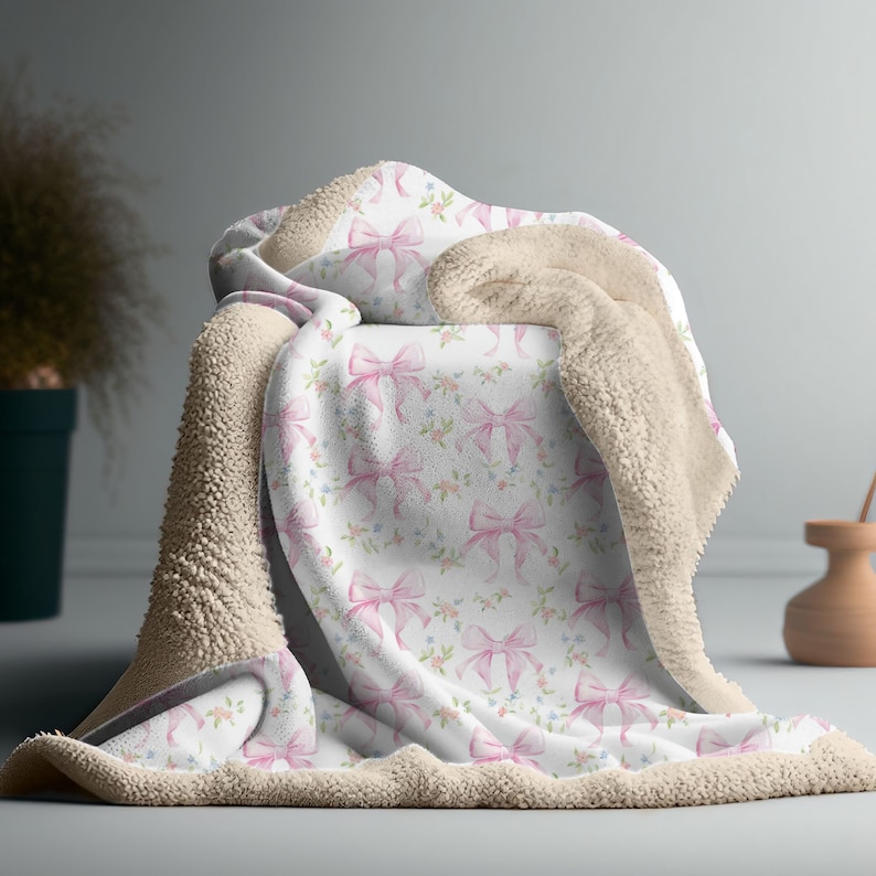 May include: A soft, plush throw blanket with a white background and a pink watercolor bow pattern. The blanket has a beige sherpa backing.