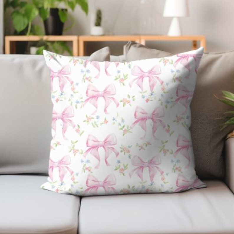 K&ouml;nnte beinhalten: Ein wei&szlig;es Kissen mit einer rosa Schleife und einem Blumenmuster. Das Kissen ist mit aquarellartigen rosa Schleifen und kleinen rosa, blauen und gr&uuml;nen Blumen verziert.