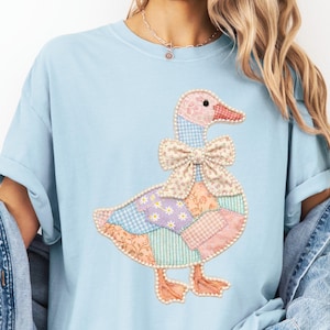 May include: Light blue t-shirt featuring a patchwork goose design. The goose has a pink and red beak, a patterned bow, and a body made of various pastel-colored fabric patches. The design is centered on the shirt.