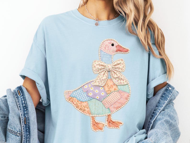 May include: Light blue t-shirt featuring a patchwork goose design. The goose has a pink and red beak, a patterned bow, and a body made of various pastel-colored fabric patches. The design is centered on the shirt.