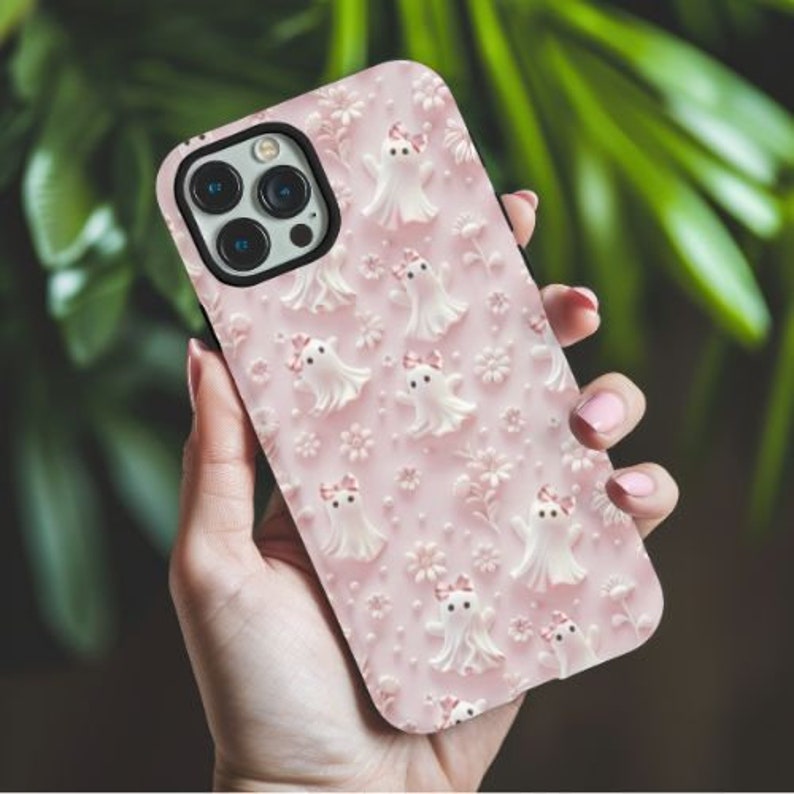 以下が含まれることがあります： 白い幽霊のパターンが施されたピンクのiPhoneケース。ケースには透明なカメラレンズと黒い縁取りがあります。