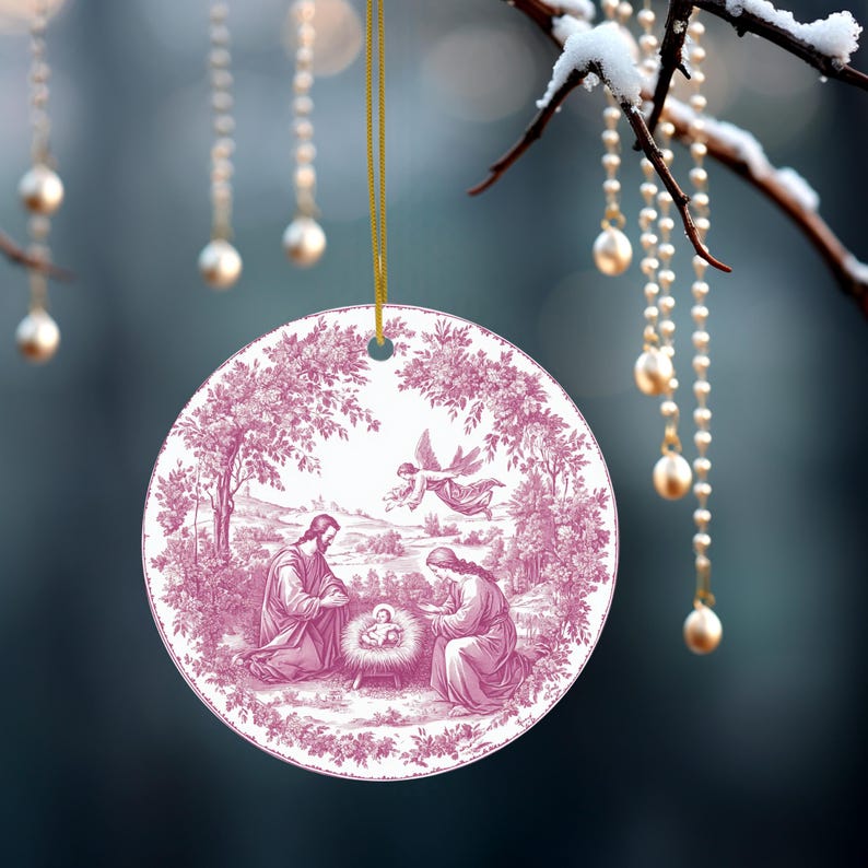 May include: A round Christmas ornament with a pink and white illustration of the Nativity scene. The ornament is hanging from a gold string. The background is blurred with strings of pearls.