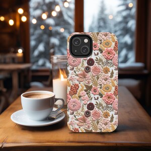 May include: A floral phone case featuring a detailed pattern of pink, peach, and brown flowers and foliage. The case is set on a wooden table next to a cup of coffee and a lit candle, creating a cozy, inviting scene.