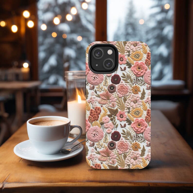 May include: A floral phone case featuring a detailed pattern of pink, peach, and brown flowers and foliage. The case is set on a wooden table next to a cup of coffee and a lit candle, creating a cozy, inviting scene.