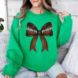 Op de afbeelding: Een groen sweatshirt met een bruine, voetbalvormige strik aan de voorkant. De strik heeft witte stiksels.