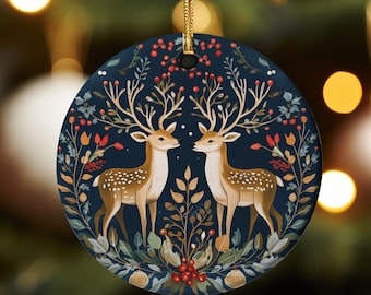 Puede incluir: Un adorno navideño redondo con fondo azul oscuro que presenta dos ciervos estilizados con cuernos, rodeados de un diseño floral con bayas rojas.