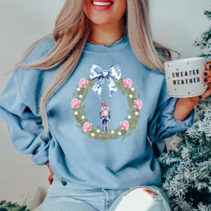 Könnte beinhalten: Ein hellblaues Sweatshirt mit einem Aquarell-Design eines Nussknackers, der in einem Kranz mit rosa Blumen und einer blauen Schleife steht. Der Text "Sweater Weather" befindet sich auf einer Tasse im Bild.