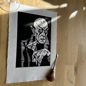 Schwarz/Weiss Linoldruck. Motiv: Nosferatu mit einem Glas Martiniin der Hand. Nosferatu trinkt diesen natürlich mit einem aufgespiessten Augapfel, anstelle einer klassischen Olive :). Der Druck ist handgezeichnet, handgedruckt und signiert.