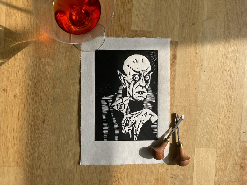 May include: Black and white woodcut print of a vampire with a sinister expression, holding a glass of wine. The print is on a white background and is framed by a white mat.