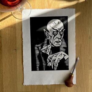 Schwarz/Weiss Linoldruck. Motiv: Nosferatu mit einem Glas Martiniin der Hand. Nosferatu trinkt diesen natürlich mit einem aufgespiessten Augapfel, anstelle einer klassischen Olive :). Der Druck ist handgezeichnet, handgedruckt und signiert.