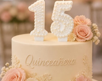 Puede incluir: Un pastel redondo de color crema decorado con flores de color rosa claro y blanco, con la palabra Quinceañera en relieve en el lateral. Dos velas blancas con forma de 15, cubiertas de pequeñas perlas blancas, están encendidas encima.