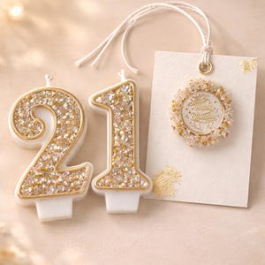 May include: A pair of white and gold birthday candles shaped as the number 21, covered in sparkling glitter. A small tag with a gold-rimmed circle that says "Happy Birthday" is also present, with a white string attached.