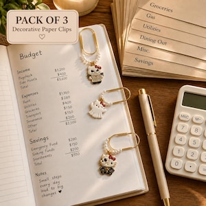 Puede incluir: Un paquete de tres clips decorativos con bucles de metal dorado, pequeñas perlas blancas y dijes de esmalte que representan personajes de gatos blancos con lazos rojos y varios atuendos, sobre un cuaderno de presupuesto abierto.