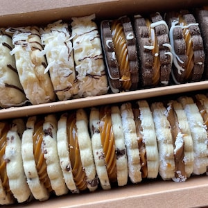 May include: A cardboard box containing two rows of sandwich cookies. The top row features coconut-topped cookies and dark chocolate cookies with caramel filling. The bottom row includes chocolate chip cookies and powdered sugar-dusted cookies.